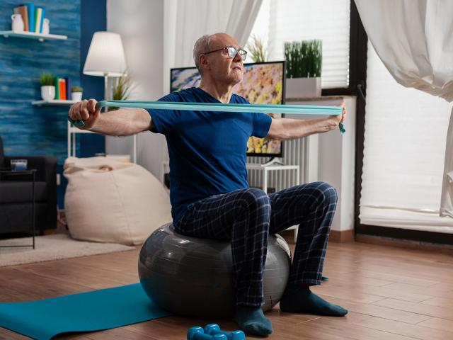 Retiree senior man sitting on swiss ball in living room doing healthcare fitness exercises streching arm muscles using resistance elastic band. Pensioner training body strength in living room