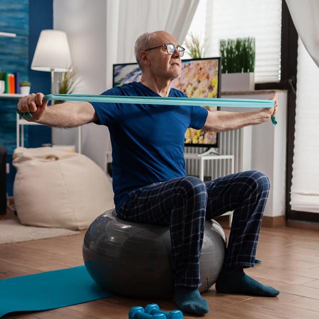 Retiree senior man sitting on swiss ball in living room doing healthcare fitness exercises streching arm muscles using resistance elastic band. Pensioner training body strength in living room