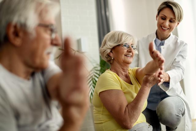 Happy senior woman and her husband doing stretching exercises with help of physical therapist at home.